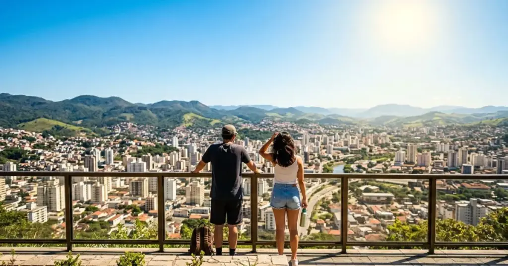 Fotografia em ângulo de primeira pessoa (POV) de duas pessoas de costas, com roupas leves e óculos de sol, paradas no Mirante do Morro do Imperador e olhando para a cidade de Juiz de Fora em um dia de sol forte e céu limpo. Tempo em Juiz de Fora