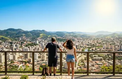 Fotografia em ângulo de primeira pessoa (POV) de duas pessoas de costas, com roupas leves e óculos de sol, paradas no Mirante do Morro do Imperador e olhando para a cidade de Juiz de Fora em um dia de sol forte e céu limpo. Tempo em Juiz de Fora