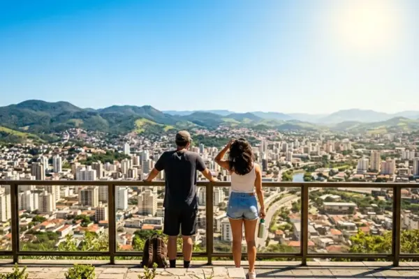 Fotografia em ângulo de primeira pessoa (POV) de duas pessoas de costas, com roupas leves e óculos de sol, paradas no Mirante do Morro do Imperador e olhando para a cidade de Juiz de Fora em um dia de sol forte e céu limpo. Tempo em Juiz de Fora